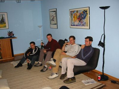 Fünf junge Männer sitzen entspannt auf Sesseln und Sofa in einem hellblauen Wohnzimmer.