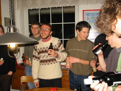 Mehrere junge Männer mit Bierflaschen stehen lachend in einem Wohnzimmer.