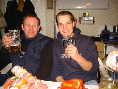 Zwei Männer prosten mit Biergläsern über einem Tisch mit Grillspießen.