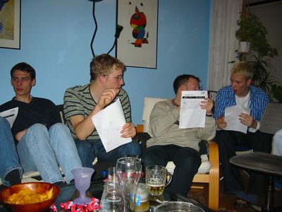 Vier junge Männer auf einem Sofa mit Zetteln in der Hand, davor Gläser.