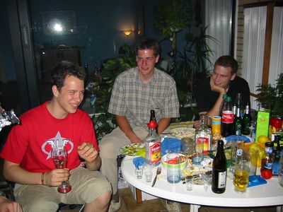 Drei junge Männer am Abend an einem vollen Tisch auf einer Terrasse.