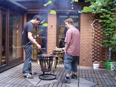 Zwei Männer stehen an einem Feuerkorb-Grill auf einer Holzterrasse.