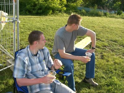 Zwei Männer sitzen mit Bechern auf einer Wiese in der Abendsonne.