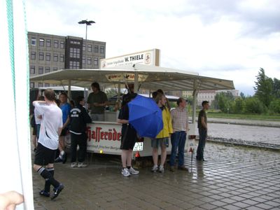 Getränkestand unter einem Dach, davor Leute, einer mit blauem Schirm.